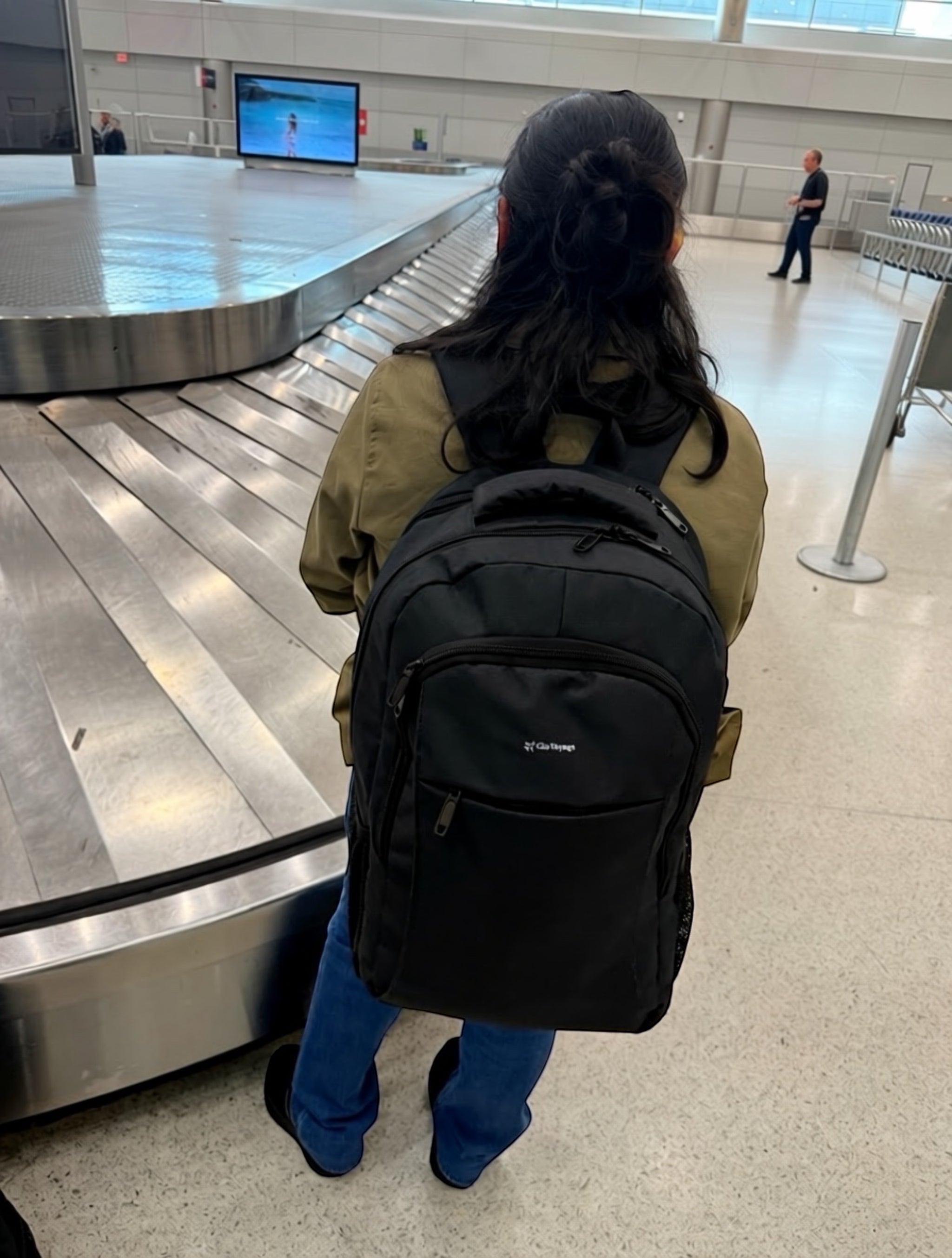 A traveler wearing a black GLOVOYAGE™ TransitPro backpack while waiting at an airport baggage claim area.